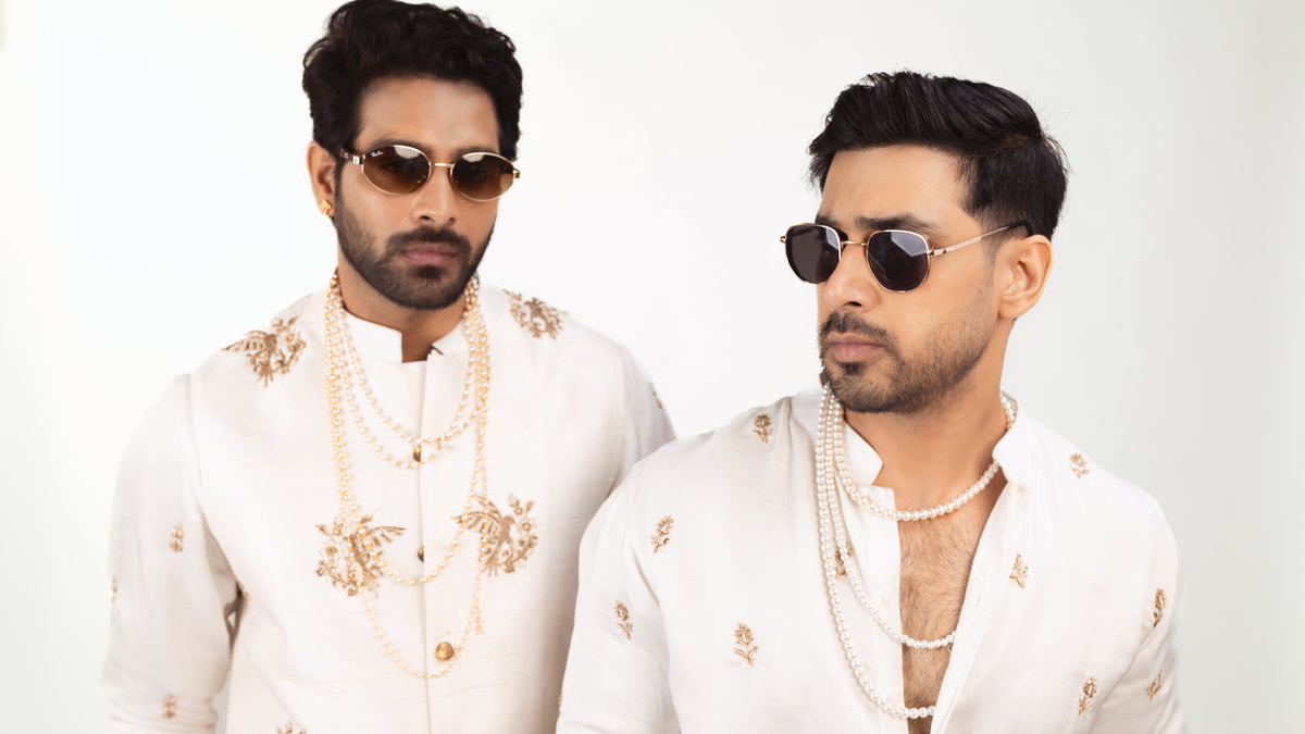 Two men wearing matching white embroidered traditional shirts with gold detailing, layered pearl necklaces, and sunglasses, standing against a plain white background.