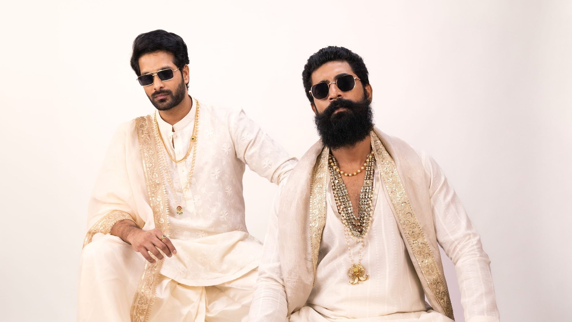 Two men in traditional white attire with gold accents sitting on a white background